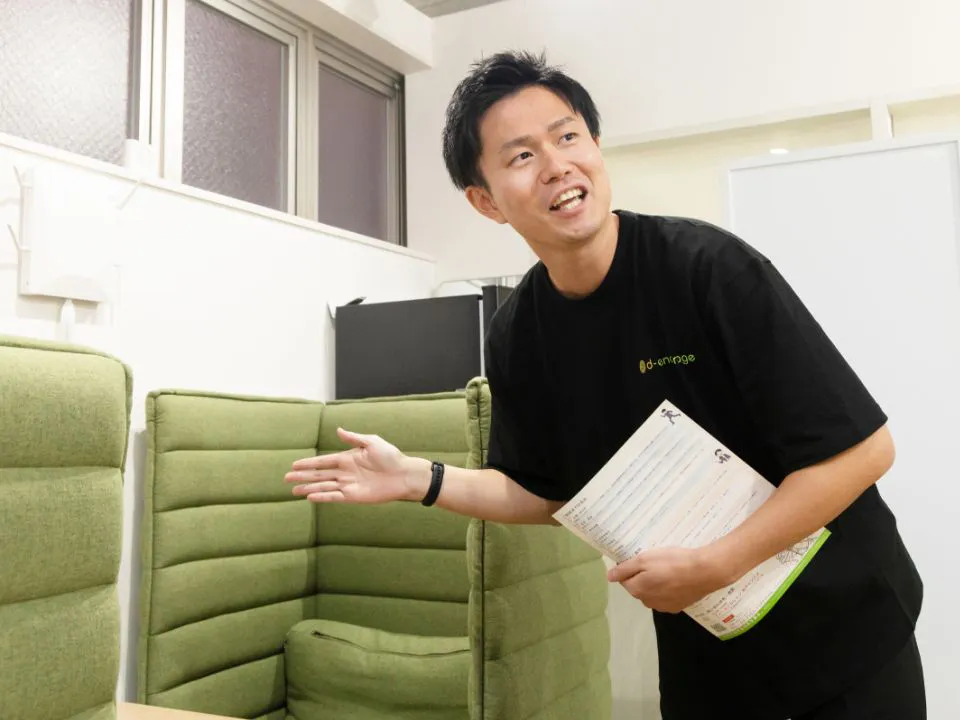 T.M.さん仕事風景。オフィスで資料を手に、ご利用者さまに緑色のソファ席を案内している様子。