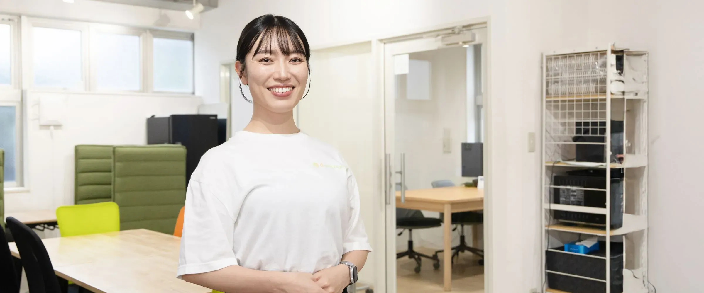 O.M.さんインタビューキービジュアル。オフィスの会議スペースで、笑顔で立っている人物の様子。背景には木製テーブルと椅子、仕切り、棚が見える。