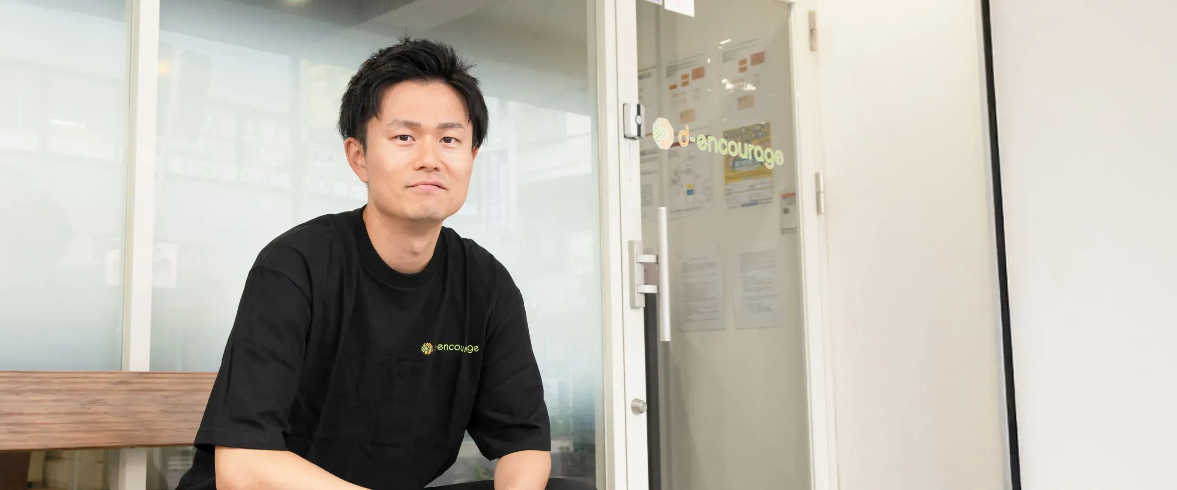 T.M.さんインタビューキービジュアル。施設の入口付近でベンチに座っている様子。