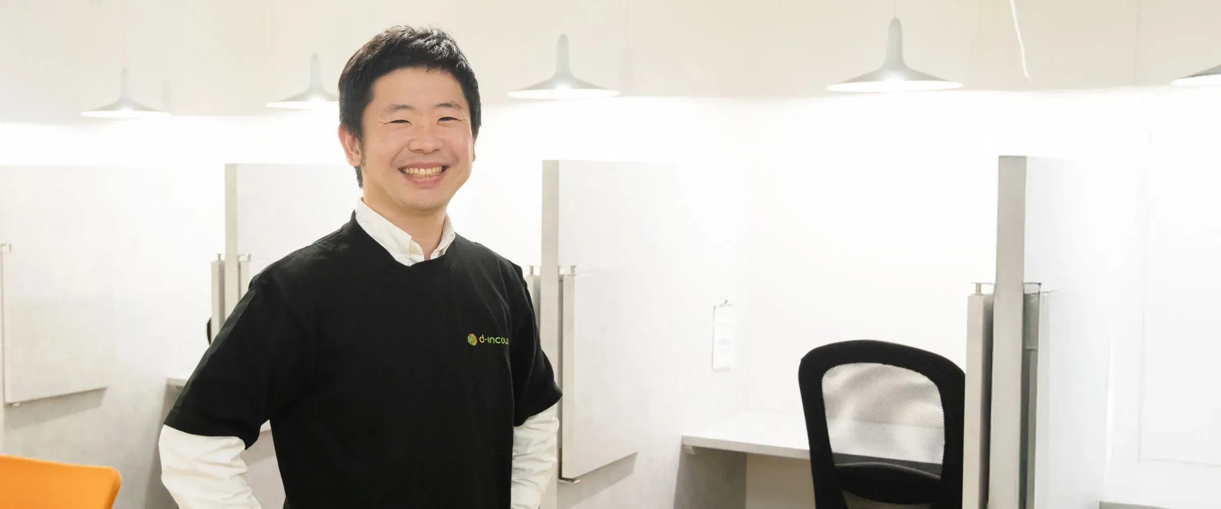 K.Y.さんインタビューキービジュアル。自立訓練施設のオフィスで、個別ブースを背景に笑顔で立っている様子。