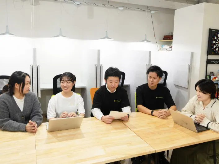 木製テーブルを囲んで複数の人がノートパソコンや資料を使って打ち合わせをしている様子。