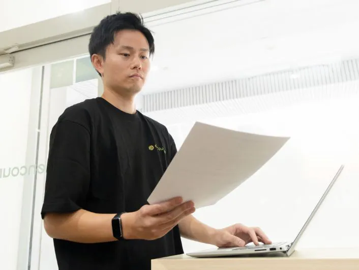 立った状態でノートパソコンを操作しながら、面談記録の書類を確認している様子。