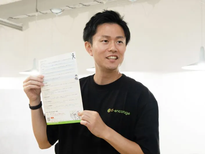 オフィスで自立訓練に関する案内資料を手に持ち、周知活動をしている様子。