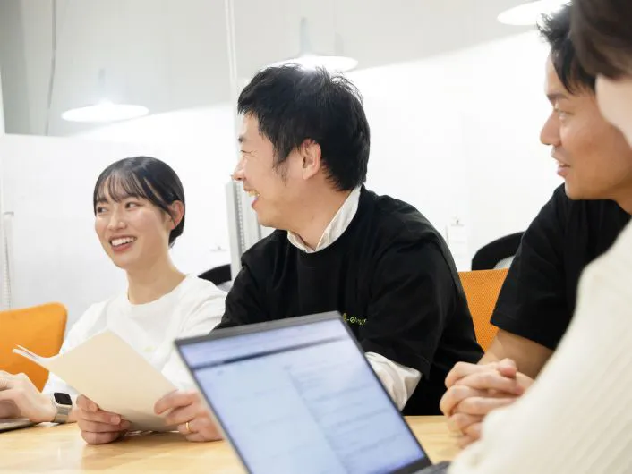 終礼の風景。複数のスタッフがテーブルを囲み、ノートパソコンや資料を使って、お互い笑顔で話し合っている様子。