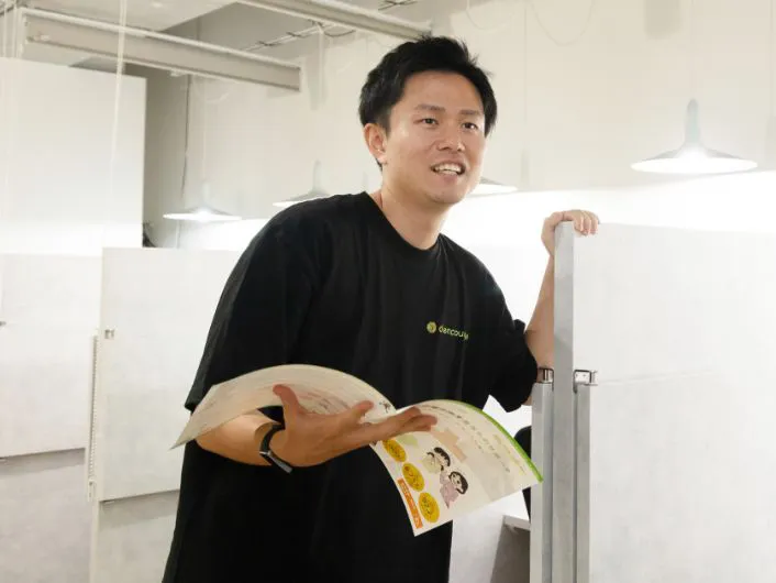 サービス管理責任者の仕事風景2。資料を手に、仕切りのあるデスクの近くに立ち、入所希望者に施設の説明をしている様子。