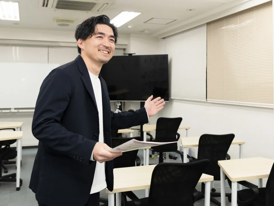 K.S.さん仕事風景。資料を持ち片手を広げてディーキャリアの訓練スペースをご案内している様子