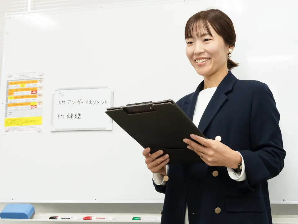 M.M.さん仕事風景。ホワイトボードの前でバインダーを持ち、プログラム内容を確認している様子