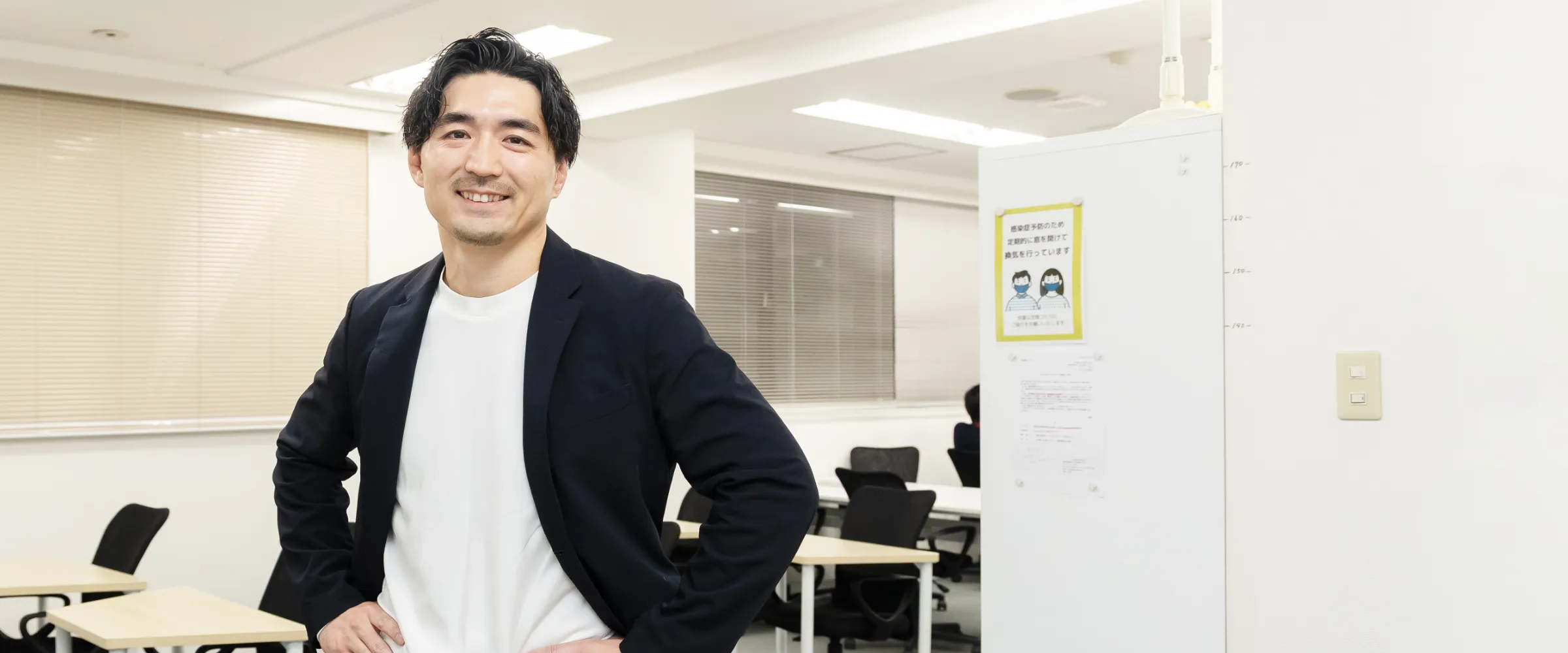 K.S.さんインタビューキービジュアル。オフィスの訓練スペースで両手を腰に置いて立っている様子