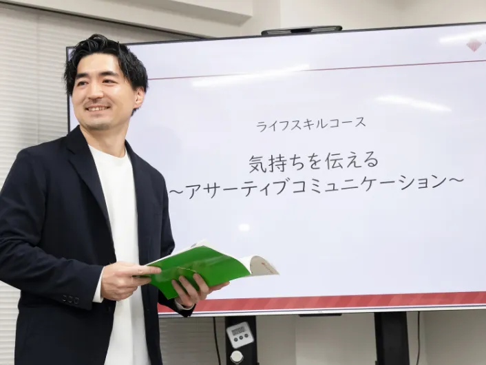 開いたテキストを片手に持ち、特大モニターの前で授業を行っている様子